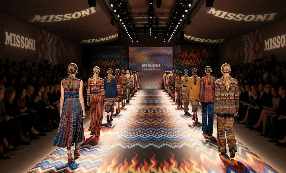 Gemini said A landscape photograph of a fashion runway show from behind the models, who are walking away from the camera. The runway, walls, and ceiling are decorated with Missoni's signature zigzag pattern in a variety of vibrant and earthy colors. The name "MISSONI" is projected onto the ceiling and walls, and on a screen at the far end of the runway, which also reads "REINVENTION". Models in patterned knit outfits walk the runway between rows of seated audience members whose faces are blurred.